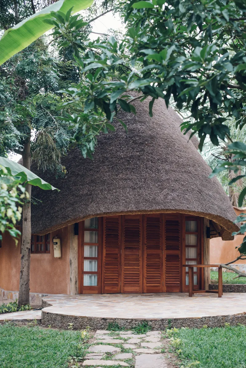 A round thatched bungalow with wooden louvred doors opening onto a circular stone terrace, framed by leaves