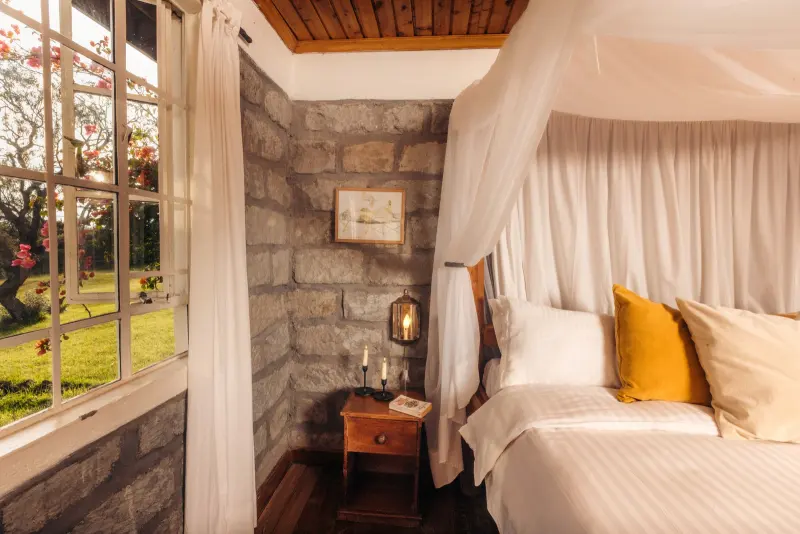 A four-poster bed hung with white netting against a stone wall, a bedside lamp lit