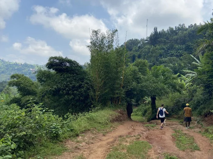 Two hikers walking a red-earth track up a green forested hillside under a bright, cloudy sky