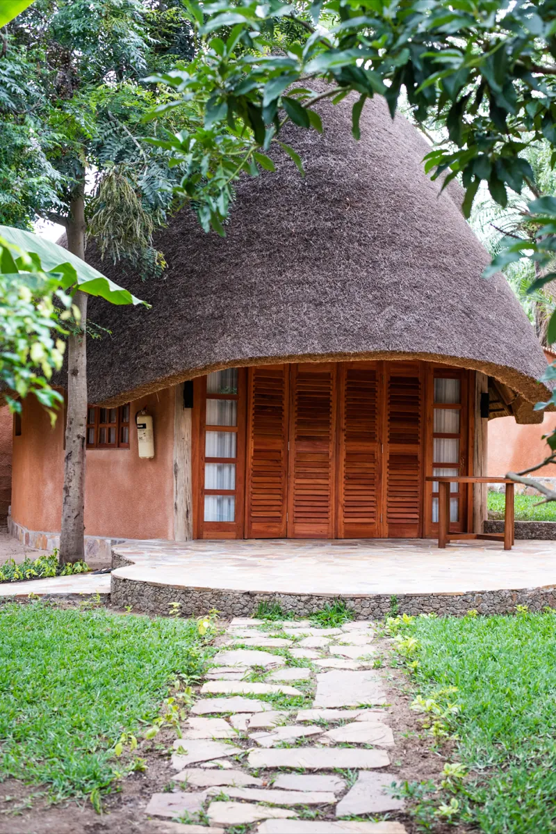 Stepping stones crossing a lawn to a round thatched bungalow with wooden louvred doors and a stone terrace