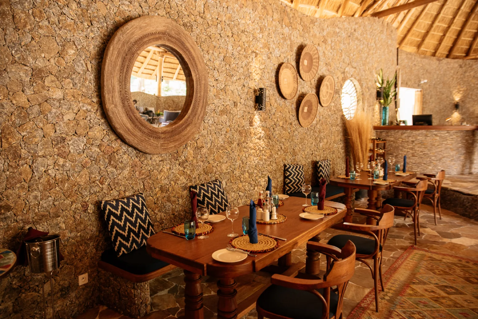 Laid wooden dining tables against a rough stone wall under the thatched roof