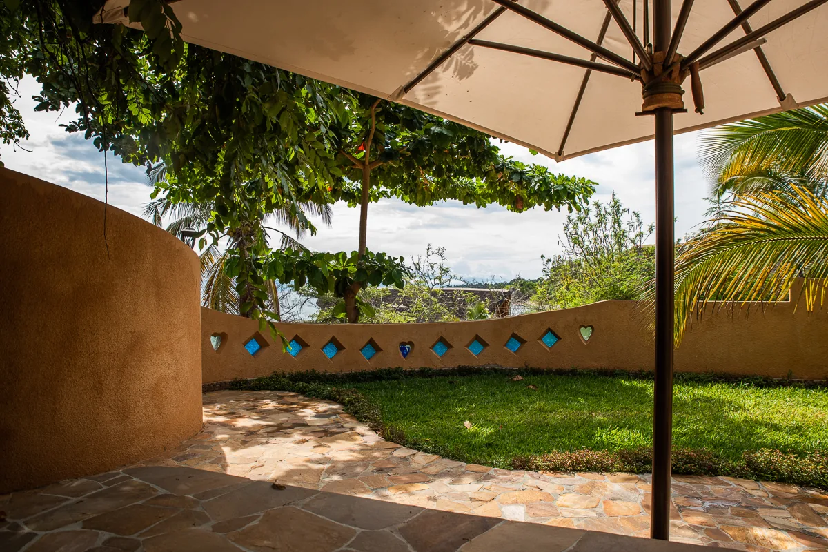 A shaded flagstone terrace under a parasol behind a curved wall inlaid with blue glass tiles