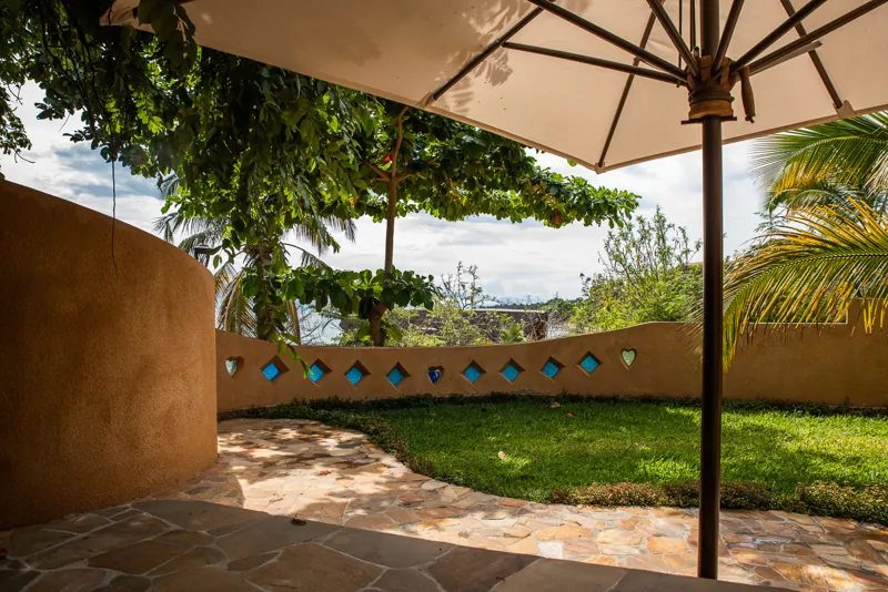 A shaded flagstone terrace under a cream parasol, looking out across the lawn to Lake Tanganyika