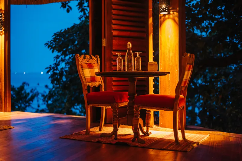 A table for two set with glasses on a timber veranda at dusk, lake and distant boat lights beyond