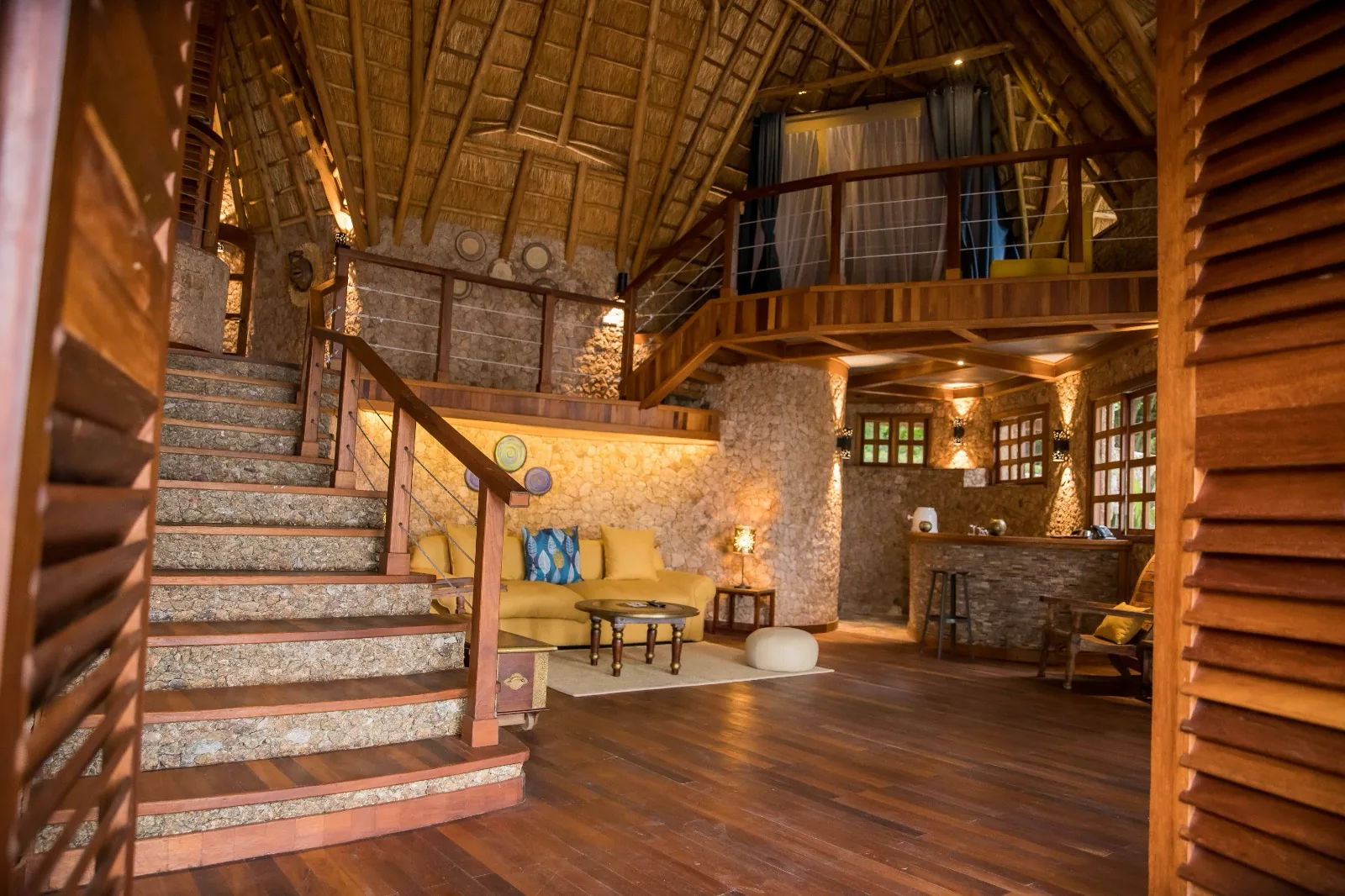 A double-height thatched living room with a yellow sofa, stone walls, staircase and mezzanine bedroom above